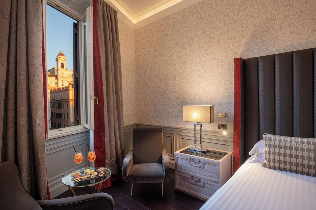A hotel room with a bed, a chair, a small table with snacks, a lamp, and a view of a building through a window.