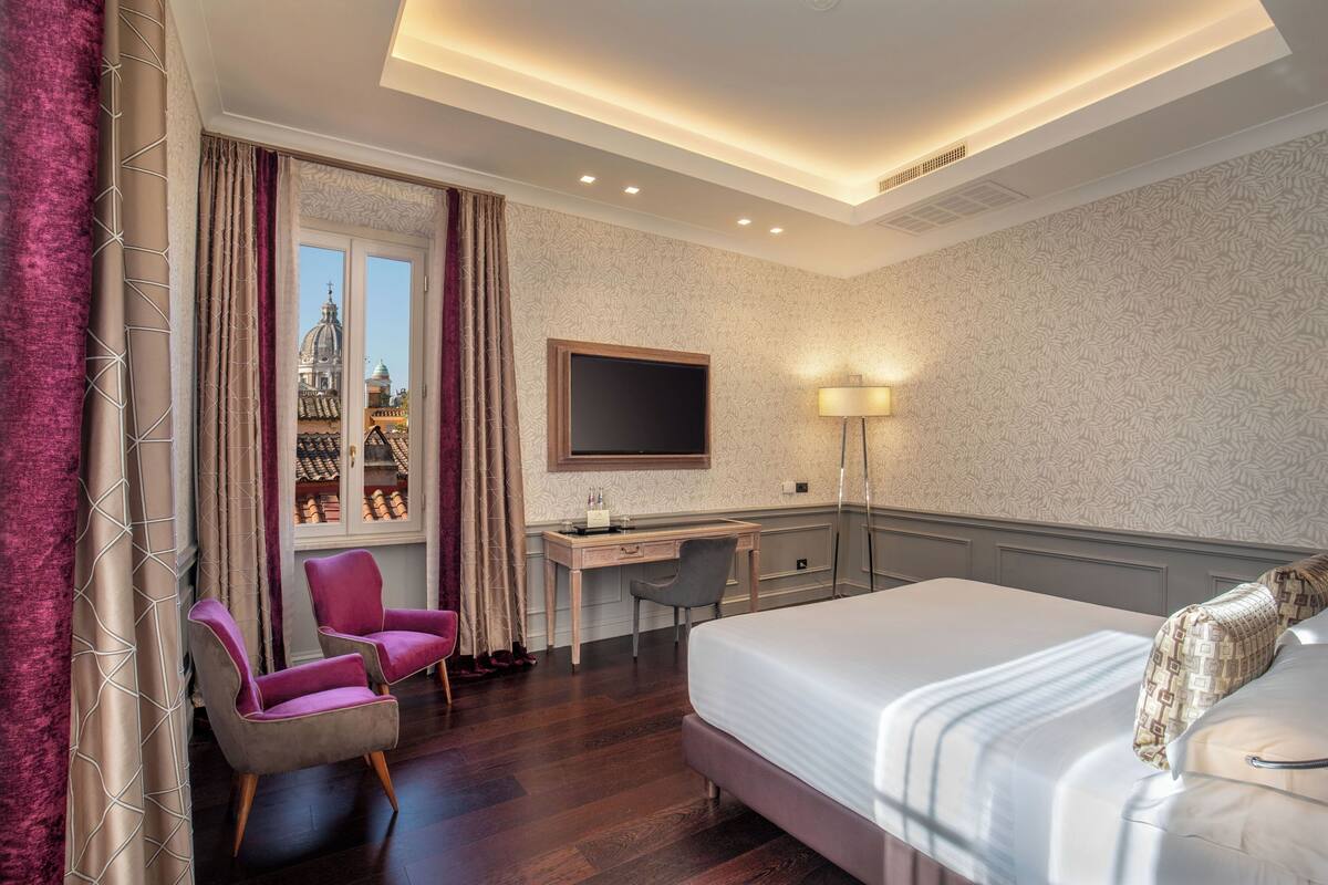 A hotel room with a bed, desk, two chairs, a TV, and a window with a view of a building.
