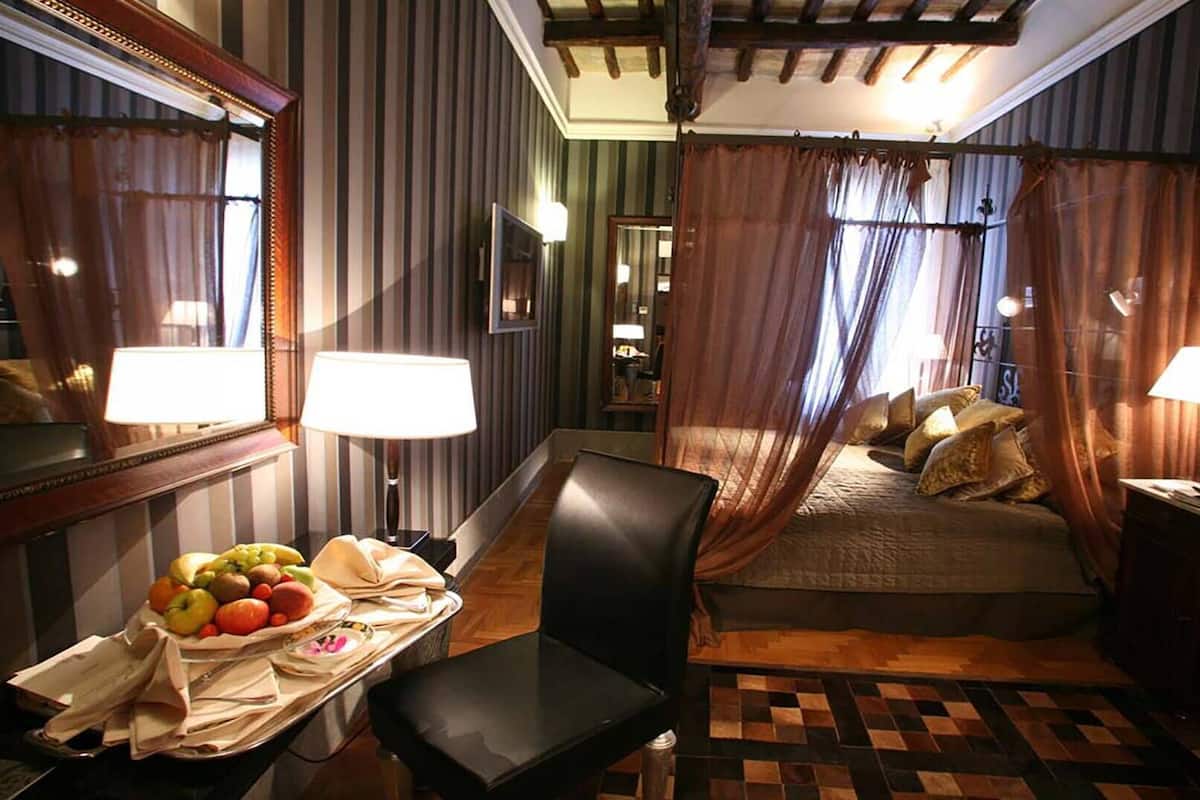 A hotel room with a bed, two bedside tables with lamps, a chair, a table with fruit and a book, and striped curtains.