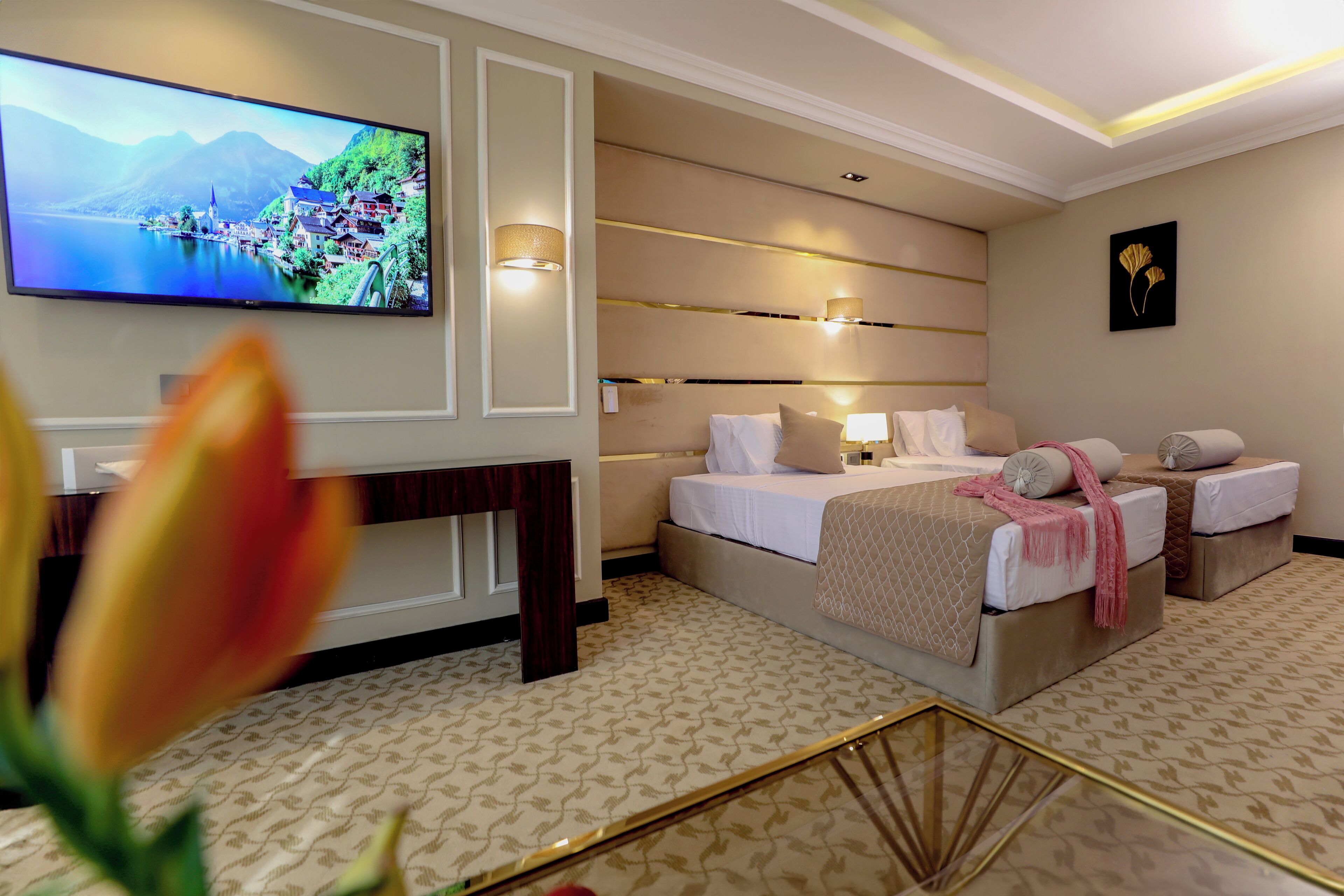 A hotel room with a large flat-screen TV, a bed with two pillows, a bedside table, and a wall-mounted lamp.