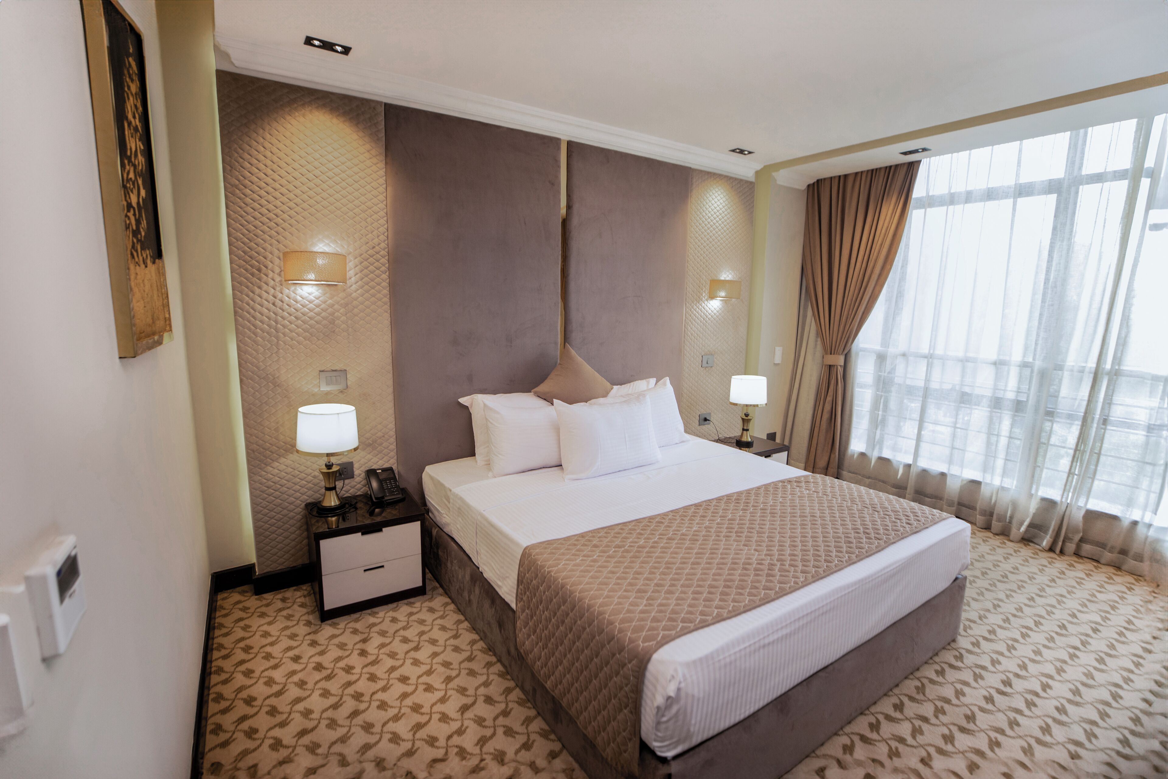 A hotel room with a large bed, bedside lamps, a nightstand, a wall-mounted phone, and a window with curtains.