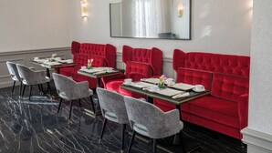 Un espace repas avec des banquettes rouges recouvertes de tissu et des chaises grises à motifs, un sol en marbre et un grand miroir au mur.