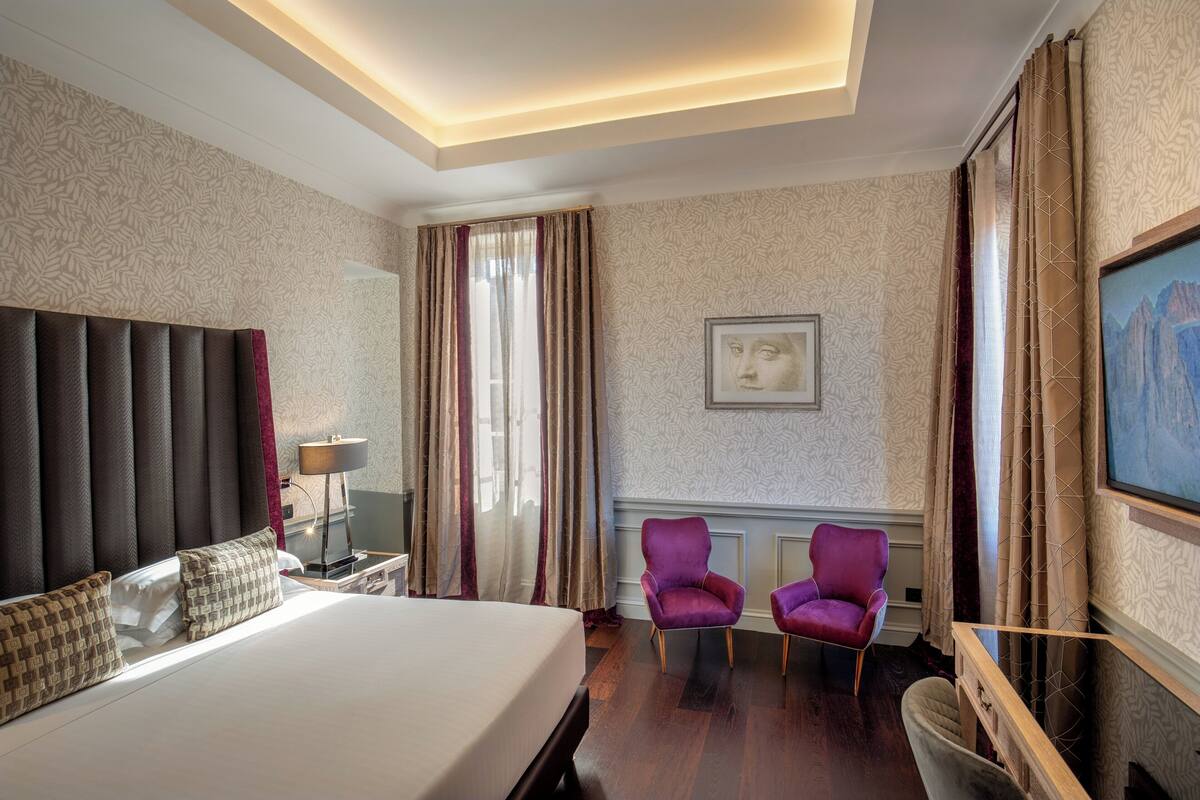 A hotel room with a large bed, two purple armchairs, a desk with a lamp, a painting on the wall, and a TV mounted on the wall.
