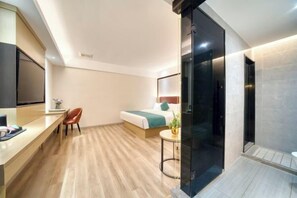 A modern hotel room with a wooden floor, a bed with a green and white bedspread, a desk with a chair, a TV, and a small table with a plant.