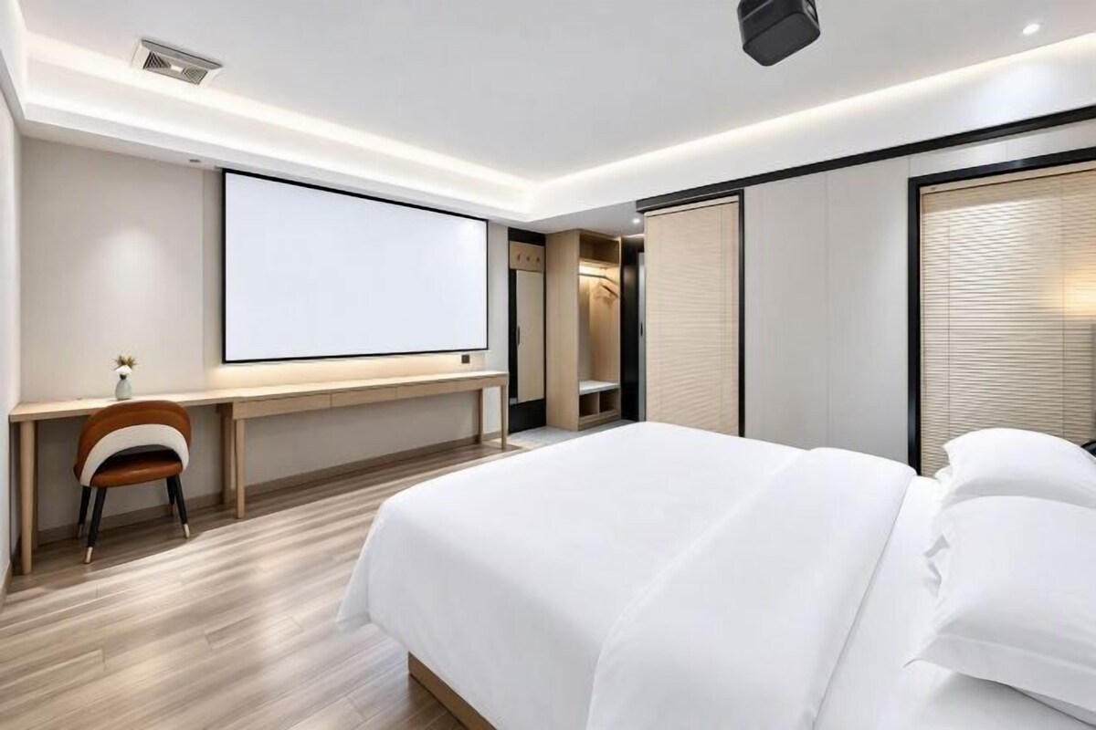 Moderni hotellihuone, jossa on suuri sänky, työpöytä tuolilla, projektorikangas ja liukuovilla varustettu vaatekaappi.