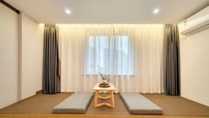 A room with a low wooden platform, two grey cushioned seating areas, a small wooden table with a vase and stones, and a window with sheer curtains. - GYA Hotel (Jiaxing Nanhu Scenic Area Nanhu Tiandi) (Jiaxing)