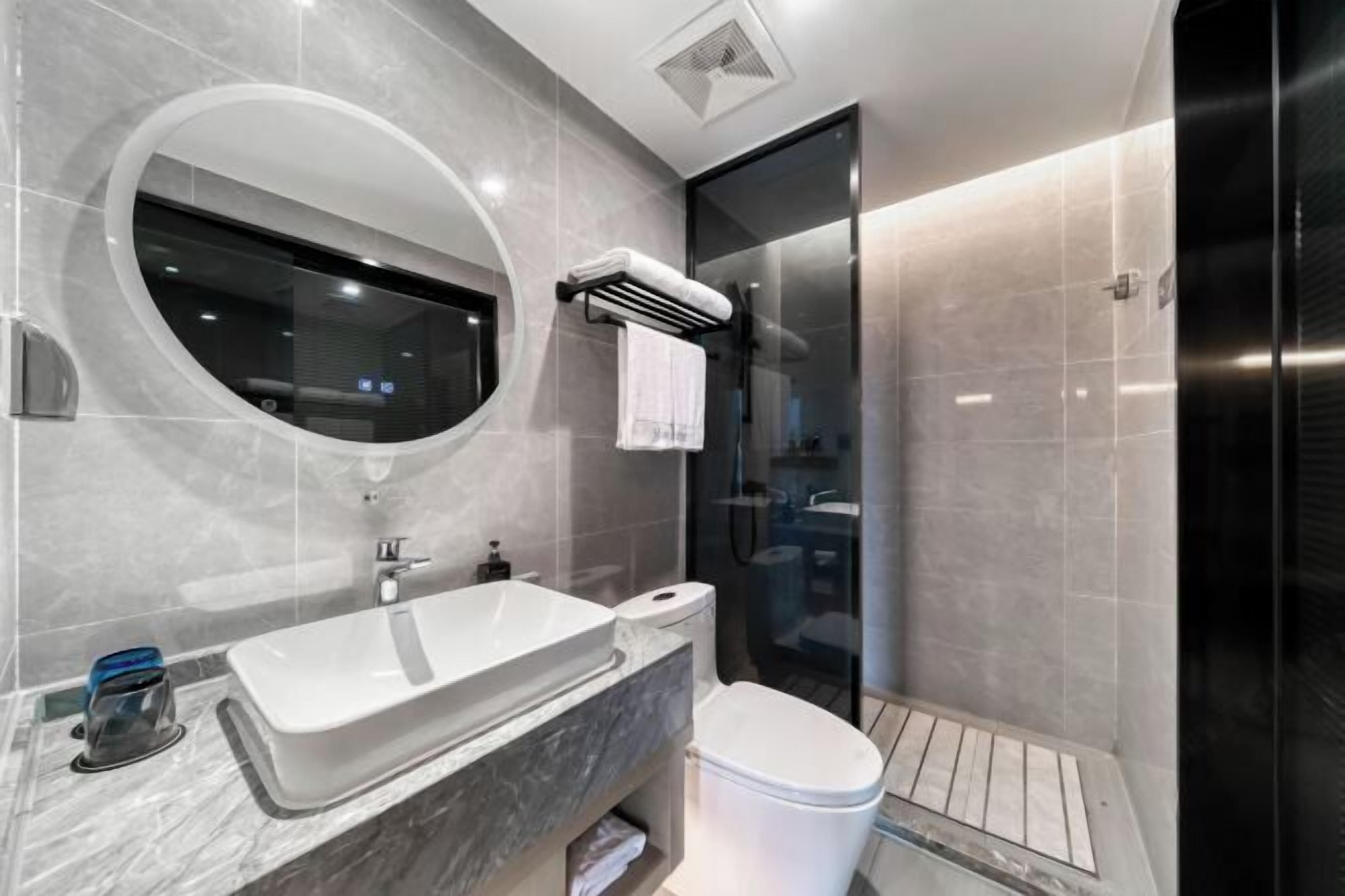 A modern bathroom with a large oval mirror, a white sink, a toilet, and a glass shower enclosure.
