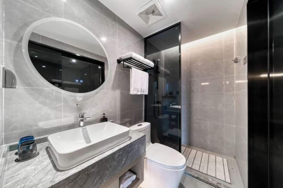 A modern bathroom with a large oval mirror, a white sink, a toilet, and a glass shower enclosure.