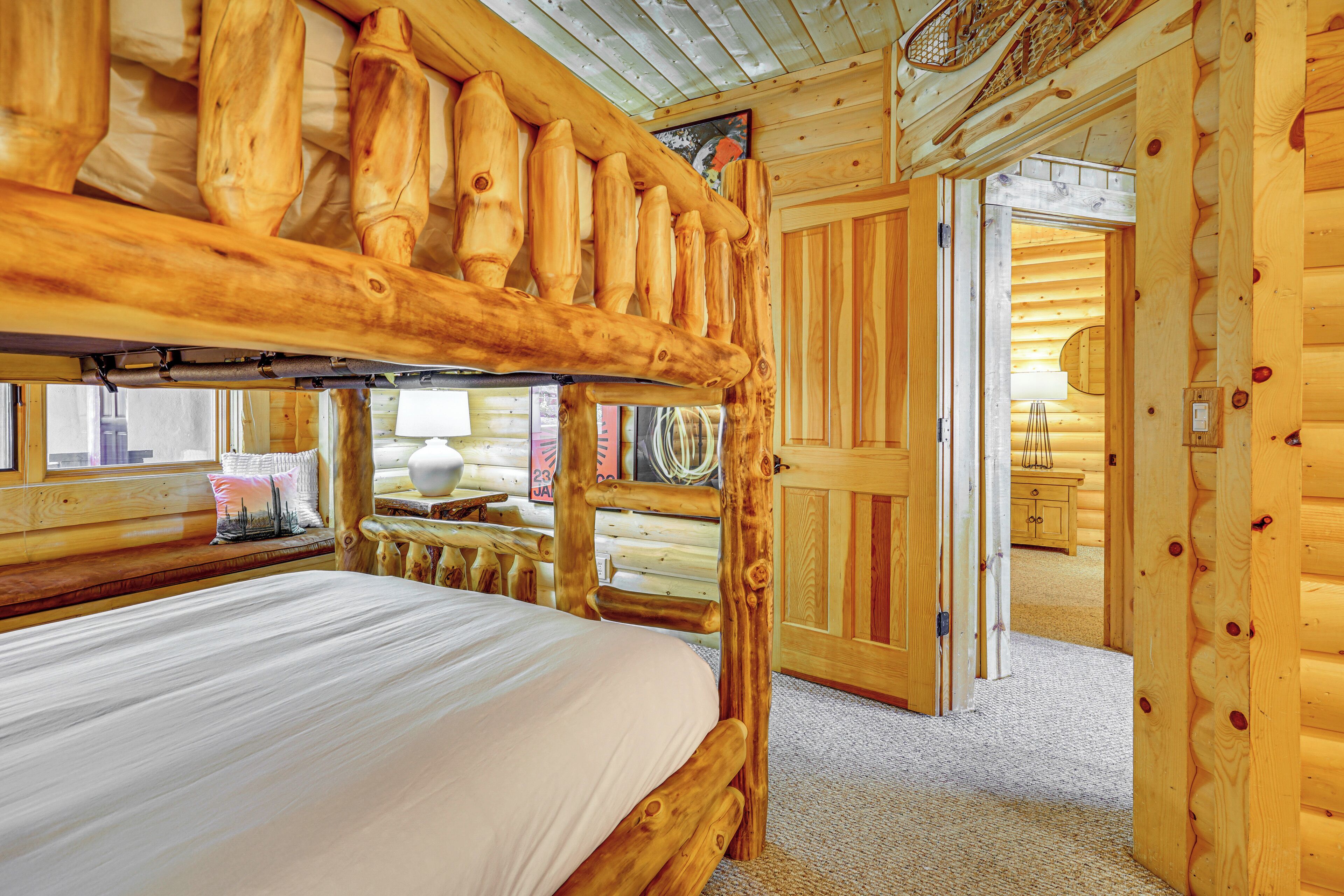 A wooden log cabin room with a bunk bed, a wooden headboard, a window with a view, and a door leading to another room.