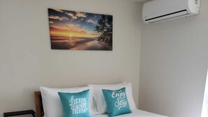 Un lit bien fait, agrémenté d’un couvre-lit à motifs, de deux coussins turquoise ornés d’inscriptions et d’un tableau représentant un coucher de soleil au-dessus.