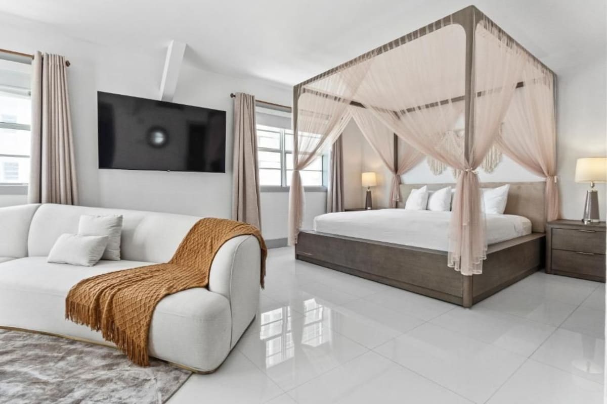 A modern bedroom with a white sofa, a four-poster bed, a flat-screen TV, and a bedside table with a lamp.