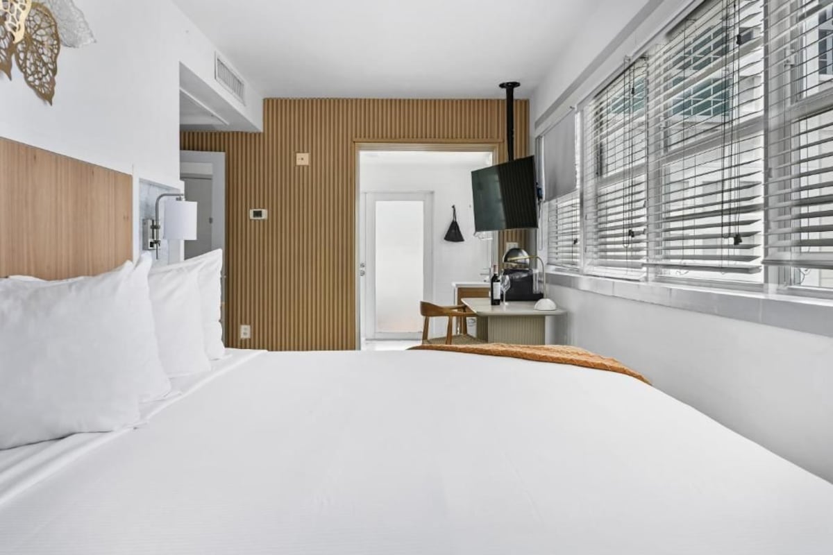 A hotel room with a bed, a desk with a chair, a TV, and a window with blinds.