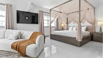 Quarto moderno com sofá branco, cama com dossel, televisão de tela plana e mesa de cabeceira com abajur.