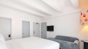 A hotel room with a bed, a grey sofa, a TV mounted on the wall, and a door with a sign.