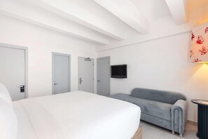 Habitación de hotel con una cama, un sofá gris, un televisor montado en la pared y una puerta con un cartel.