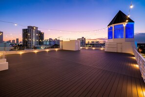 Terraza en la azotea con vistas al horizonte de la ciudad, luces cálidas y una estructura pequeña con ventanas de tono azulado.