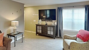 Una sala de estar con una televisión de pantalla plana, un sofá, un sillón, una mesita con una lámpara y un aparador.