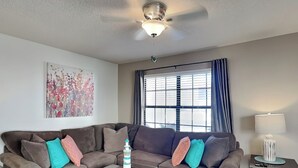 Una sala de estar con un sofá modular, una mesa de centro, un ventilador de techo, una ventana con persianas y una lámpara sobre una mesita auxiliar.