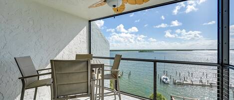 A balcony with a table and chairs, a ceiling fan, and a view of a body of water with boats and docks.