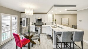 Una cocina moderna con armarios blancos, una isla de mármol y un comedor con mesa y sillas de vidrio.