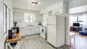 Una cocina moderna con armarios blancos, horno empotrado y refrigerador. Hay una mesa de madera con cafetera y una computadora portátil.