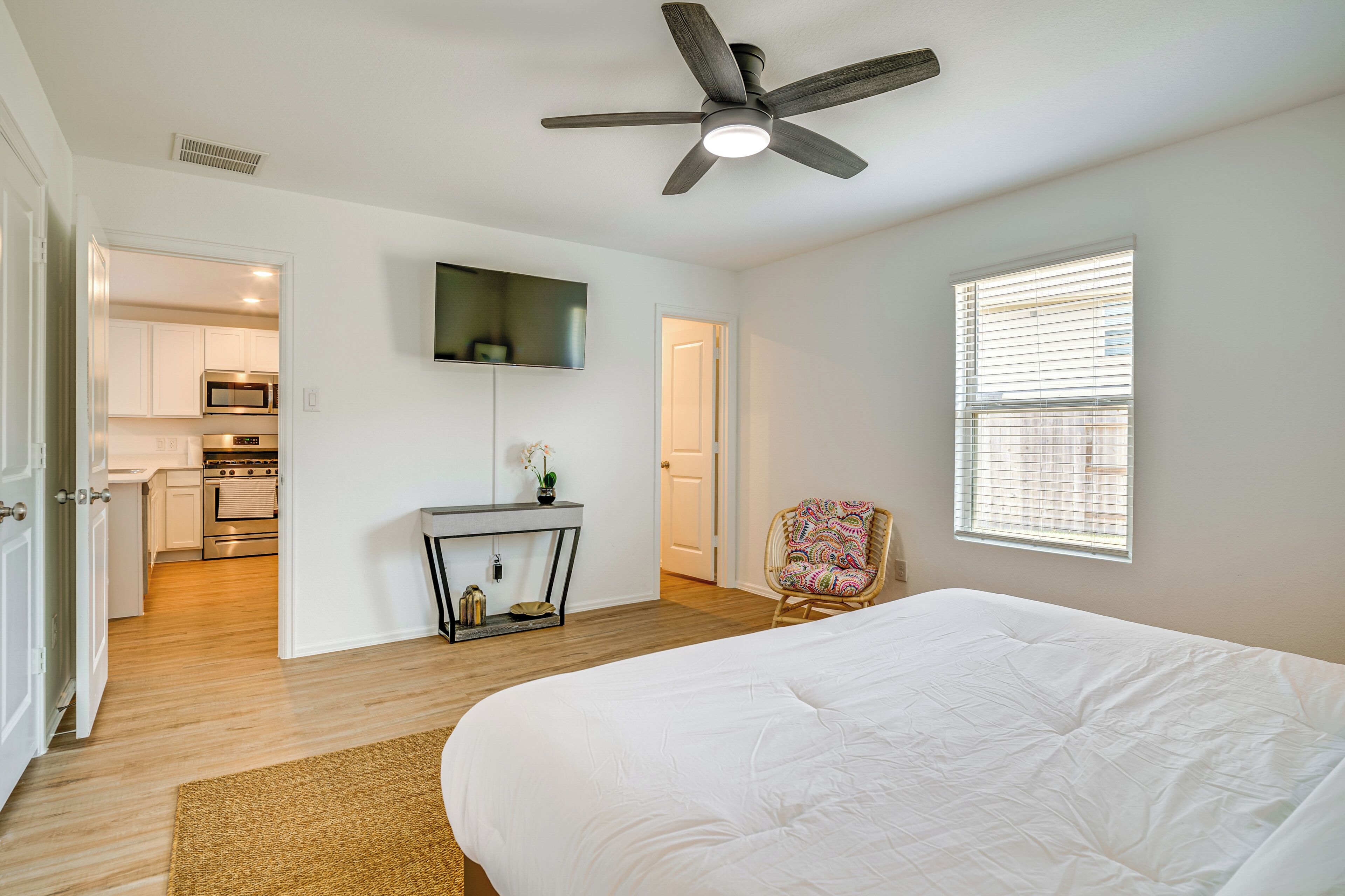 A bedroom with a bed, a ceiling fan, a TV mounted on the wall, a chair, and a window with blinds.