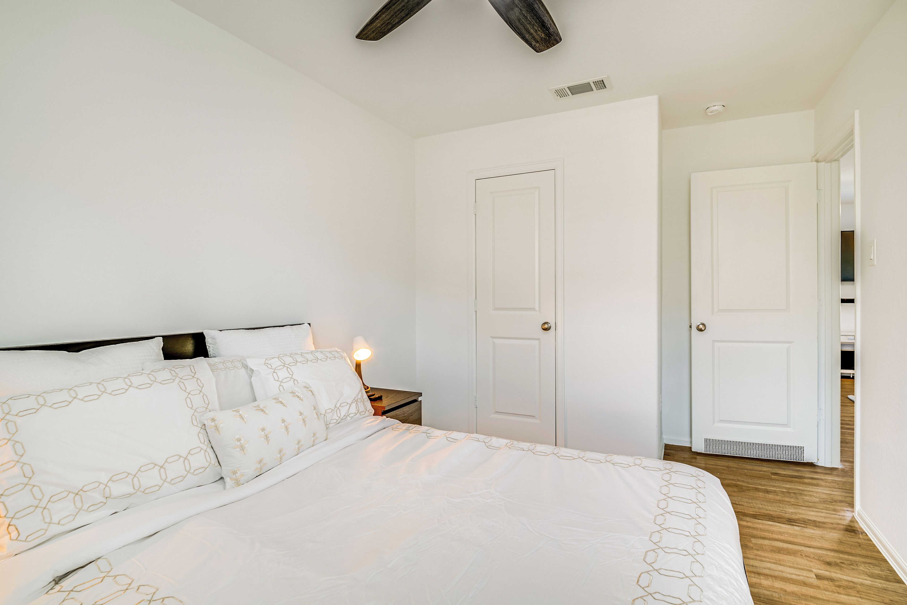 Ein Schlafzimmer mit einem großen Bett, weißen Wänden und Holzboden. Zwei weiße Türen und ein Nachttisch mit Lampe sind vorhanden.