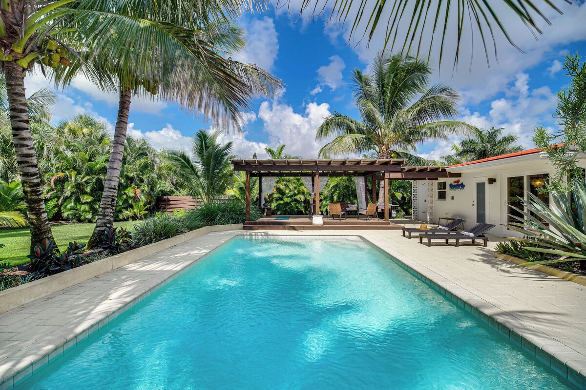 A swimming pool with lounge chairs, a pergola, and palm trees.
