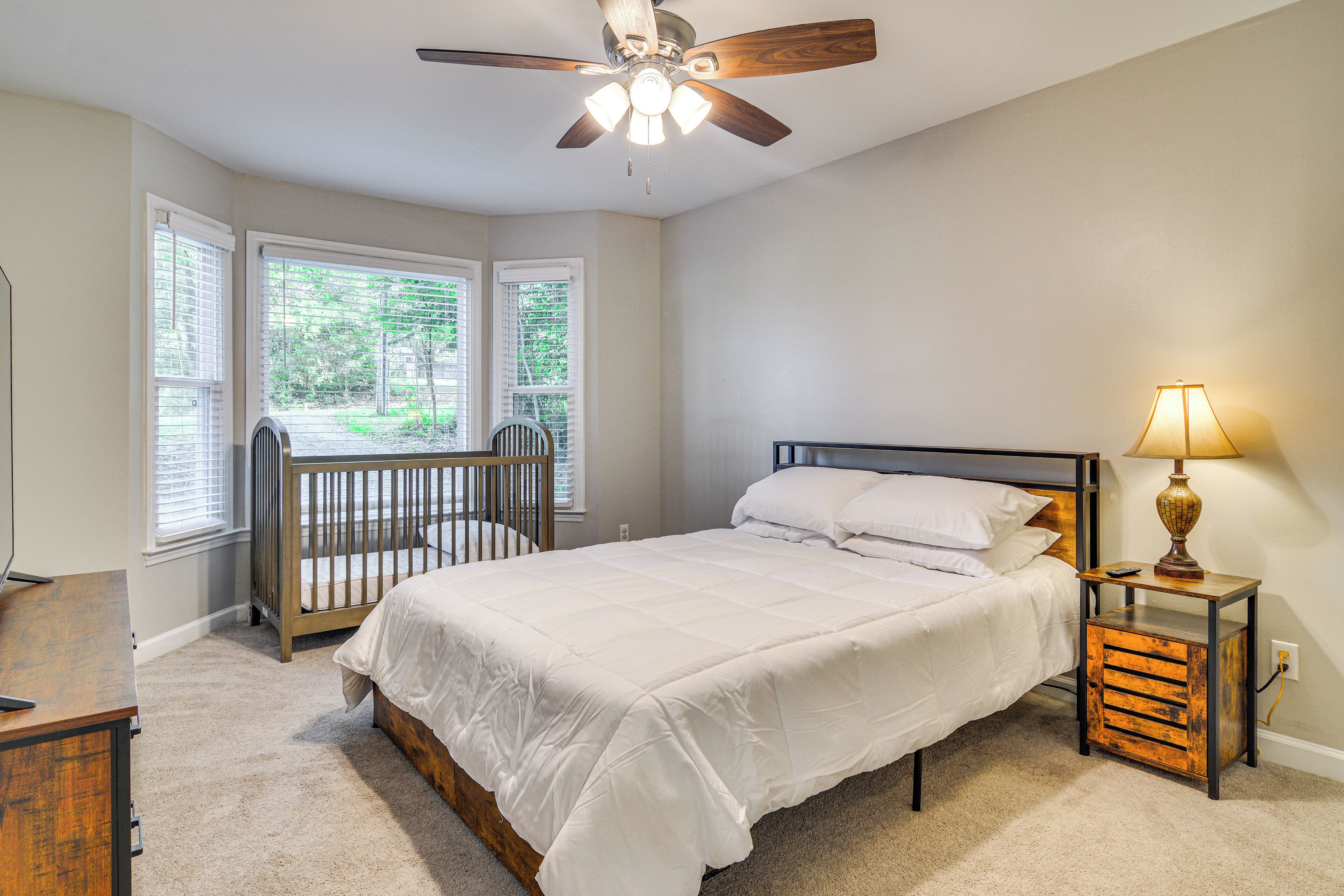 A bedroom with a bed, a nightstand with a lamp, a crib, and a ceiling fan.