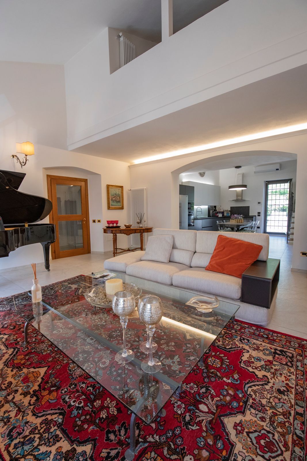 A spacious living room with a grand piano, a glass coffee table with wine glasses, a patterned rug, and a modern kitchen in the background.