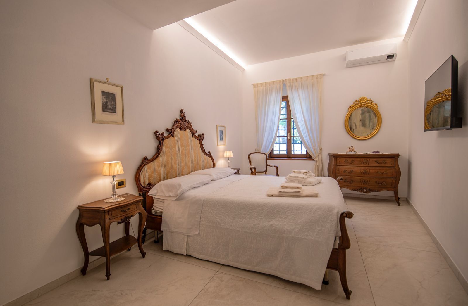 Un dormitorio con una cama grande, un aparador de madera, una silla, una mesita de noche con una lámpara, una ventana con cortinas y un cuadro en la pared.