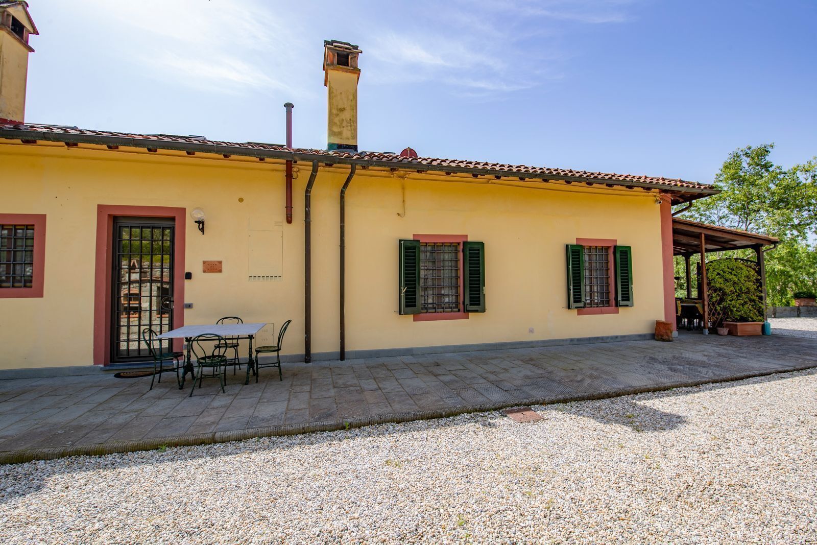 A yellow building with a tiled roof, a door with a metal gate, and two windows with green shutters.