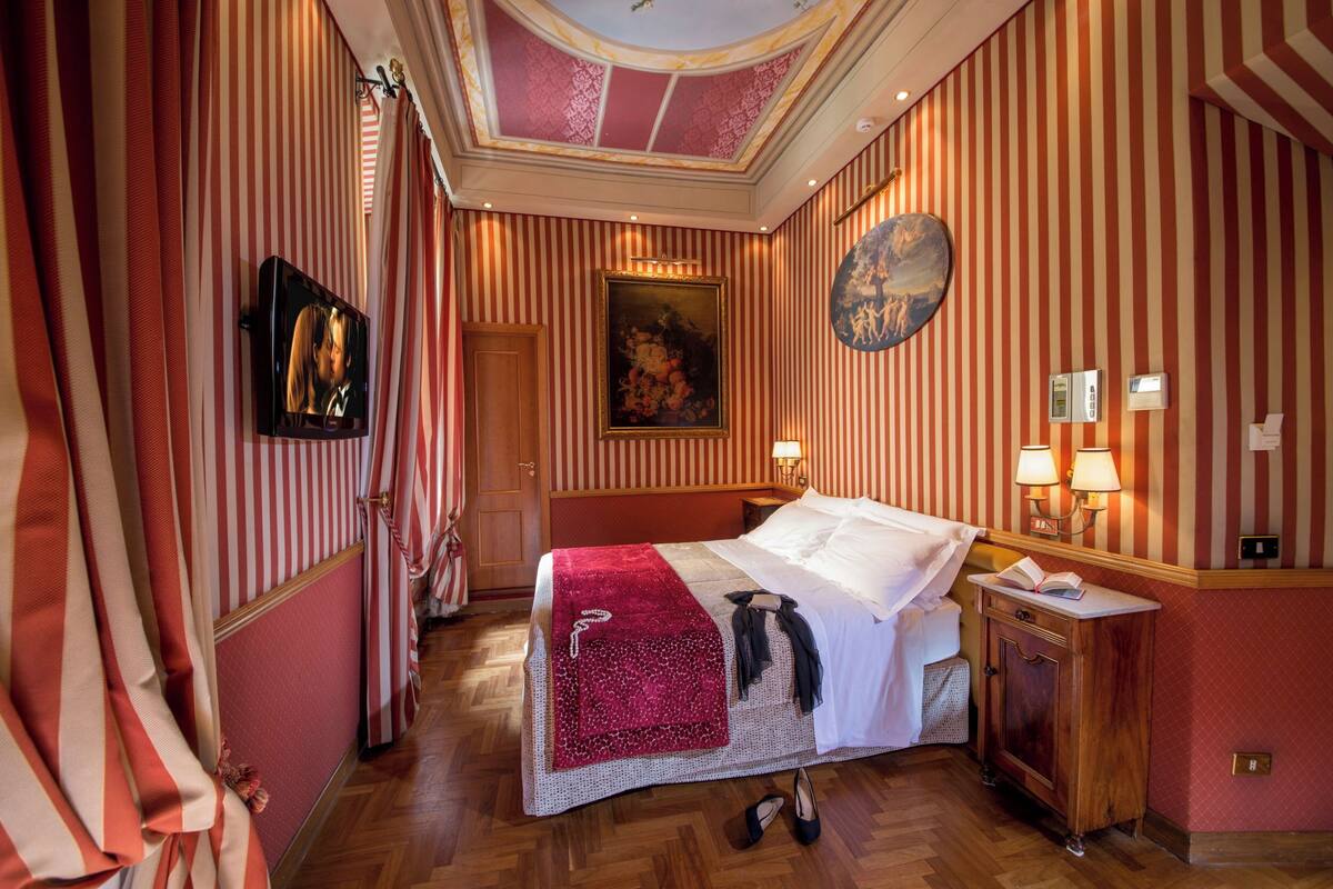 Una habitación de hotel con papel tapiz a rayas, una cama con cubrecama blanco, una manta roja, un cuadro en la pared y un reloj.