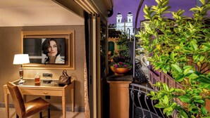 A hotel room with a desk, chair, lamp, and a television. There is a balcony with plants and a view of a building with a clock tower. - The Inn at the Spanish Steps Exclusive Collection - Roberto Di Rienzo (Rome)