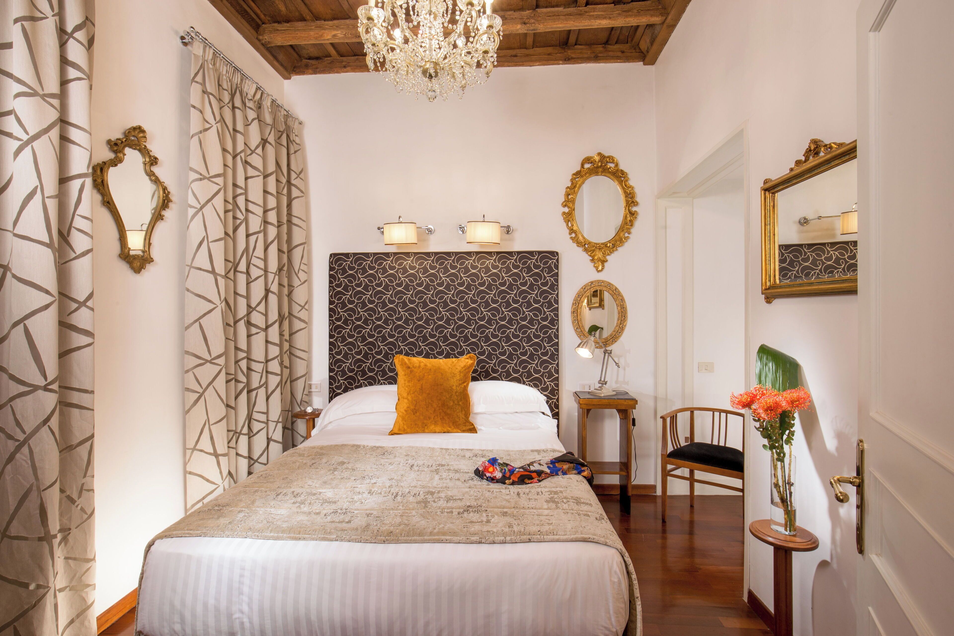 A hotel room with a large bed, a chandelier, a mirror, and a small table with a vase of flowers.