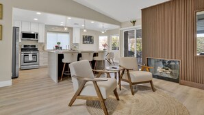 A modern kitchen with white cabinets, a microwave, and a built-in oven. There are two white chairs and a small table in the living area.