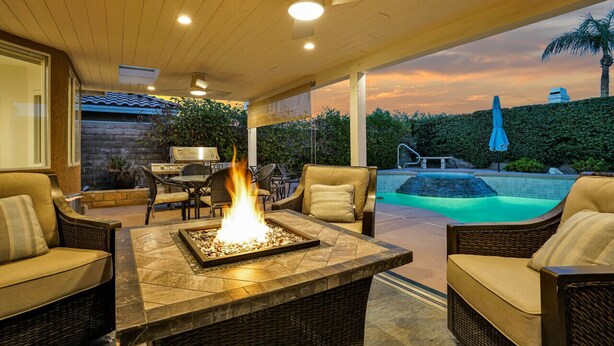 A covered outdoor patio with a fire feature, wicker furniture, and a view of a swimming pool at sunset.