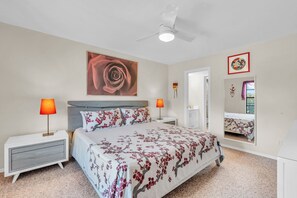 A bedroom with a bed, nightstands, a ceiling fan, a large framed artwork, a mirror, and a door.