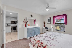 A bedroom with a bed, dresser, desk, and a view of the kitchen.