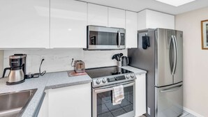A modern kitchen with white cabinets, a stainless steel refrigerator, a built-in microwave, a stovetop, and a coffee maker. - H-247 - Second Floor With Lake and Fountain View (Naples)