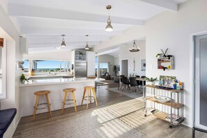 A modern kitchen with a central island, bar stools, a dining area with chairs, and a view through large windows. - Beachfront Retreat Su (Mount Maunganui)