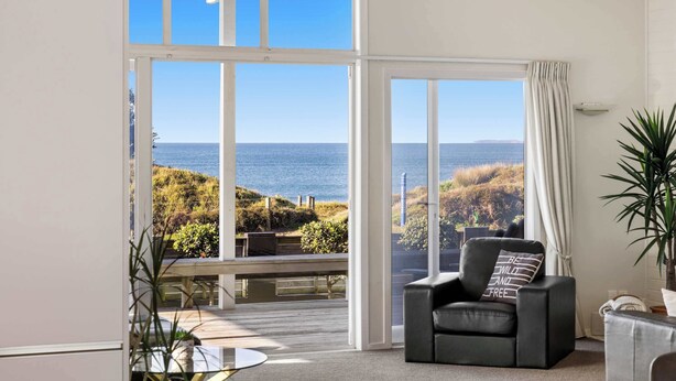 A spacious room with a large sliding glass door leading to a balcony. The room has a comfortable seating area with a black leather chair and a small table. There is a potted plant on the right side of the room. - Beachfront Retreat Su (Mount Maunganui)