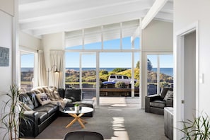 A spacious living room with a large glass door leading to a scenic outdoor view, a black leather sofa, a round coffee table, and a view of a parked vehicle.