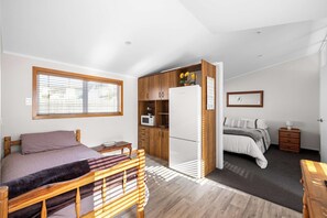 A bedroom with a bed, bedside table, and a small wooden desk. There is a kitchenette with a microwave and a refrigerator. A window provides natural light.