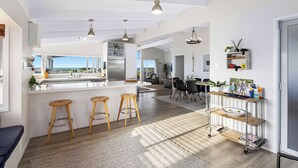 A modern kitchen with a central island, bar stools, a dining area with chairs, and a view through large windows. - Beachfront Retreat Su (Mount Maunganui)