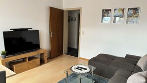 A modern living room with a flat-screen TV on a wooden stand, a glass coffee table with remote controls, a grey sofa, and a wooden door leading to another room. - Vacation apartment by the vineyards, great wine hiking trails nearby (Öhringen)