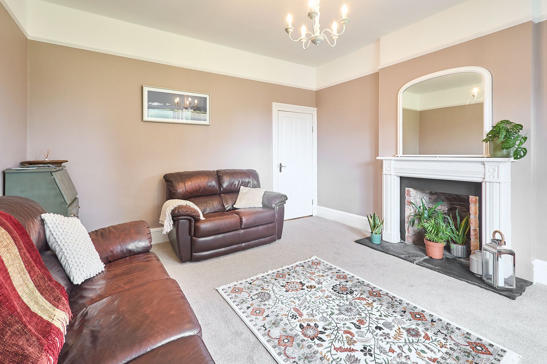 A living room with a brown leather sofa, a white door, a fireplace with plants, and a chandelier.