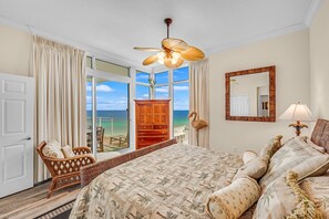 A bedroom with a large bed, a ceiling fan, a wooden wardrobe, a wicker chair, and a view of the ocean through a sliding glass door.