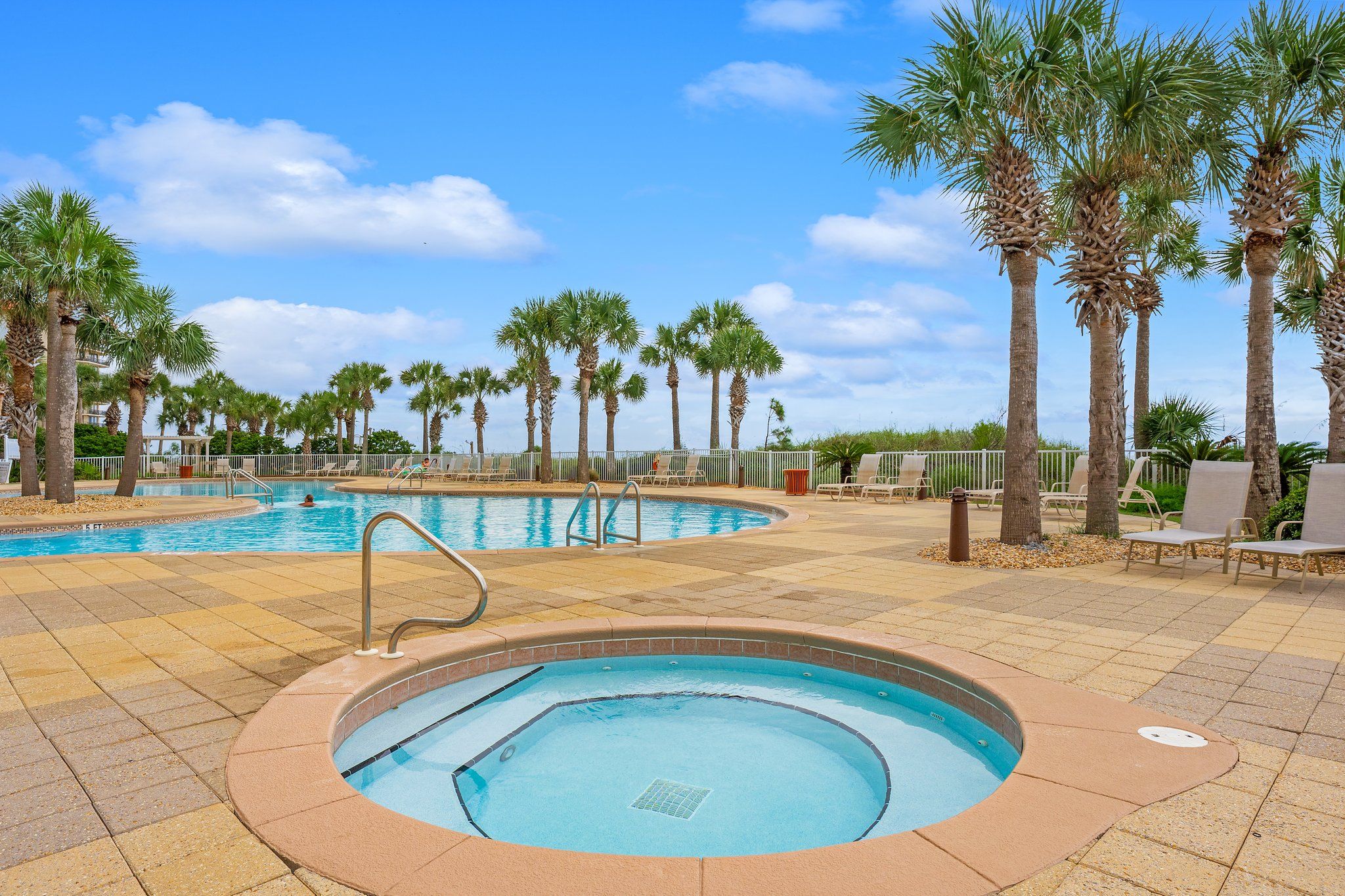A swimming pool area with a hot tub, palm trees, and lounge chairs.