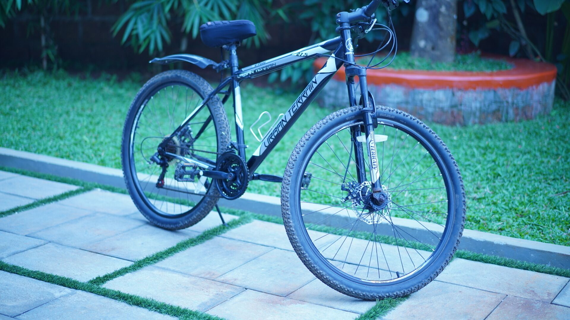 A bicycle parked on a paved path with grass and a planter in the background.
